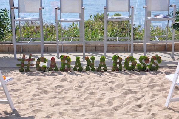 Large botanical hashtag sign reading #cabanaboss by flowerduet.com sitting on sand in front of Pacific Ocean